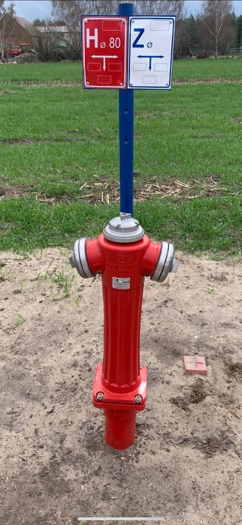Na zdjęciu przedstawiony jest czerwony hydrant. Za hydrantem stoi 
niebieska rura z dwoma tabliczkami jedna w kolorze czerwonym druga w 
kolorze białym. Hydrant stoi na nieutwardzonej drodze. W oddali widać 
pole.
