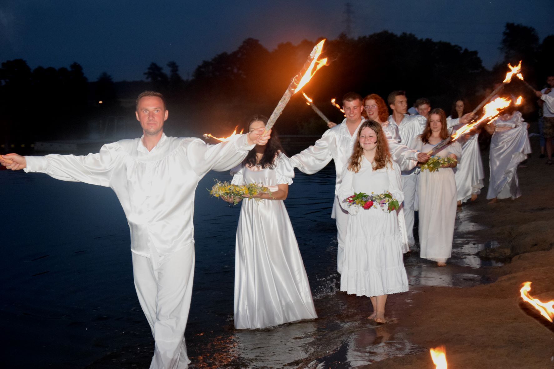 Ujęcie z obchodów Nocy Świętojańskiej w Gołanicach. Mężczyźni, kobiety oraz dzieci ubrane na biało. Mężczyźni trzymają pochodnie a kobiety wianki spacerując przy brzegu jeziora.&nbsp;