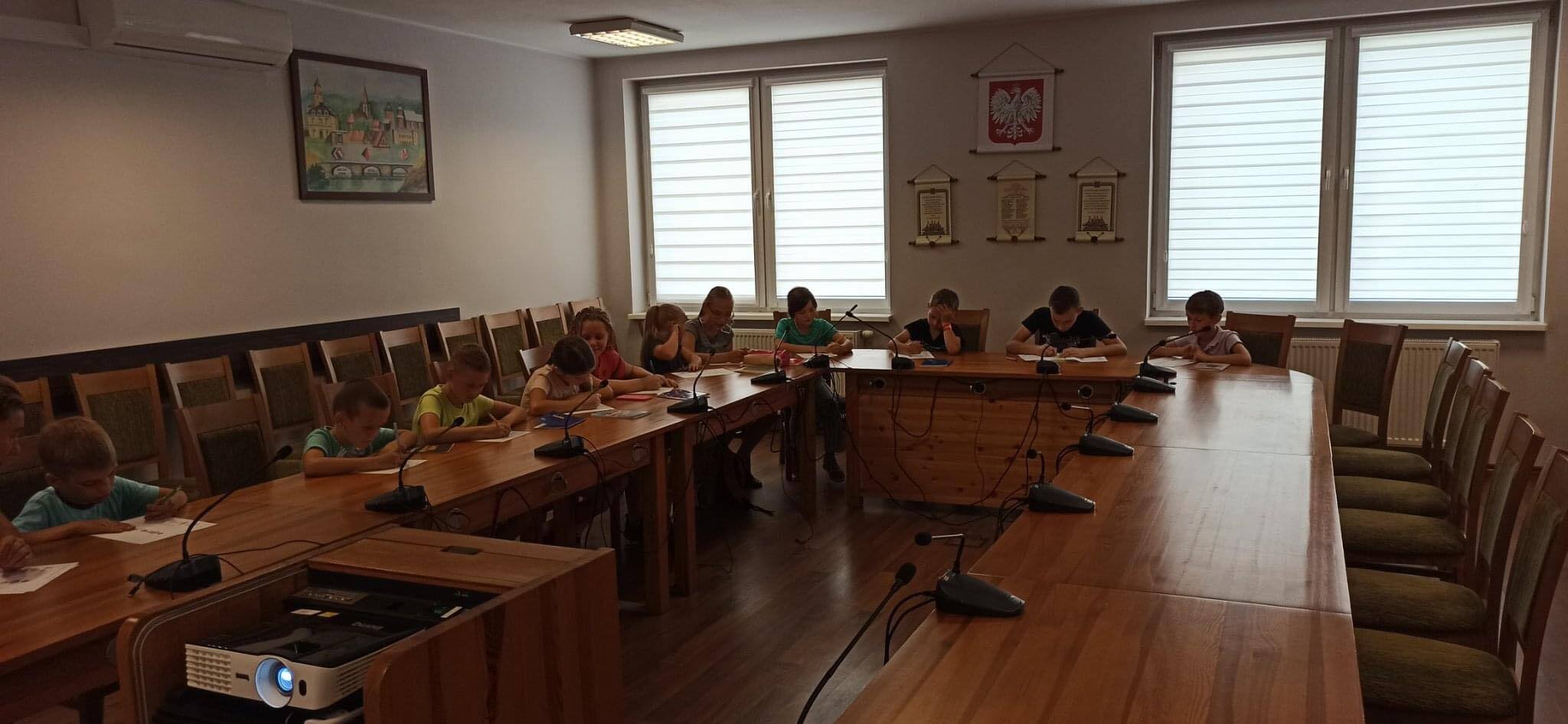 Fotografia przedstawia 11 siedzących przy stole dzieci rozwiązujących zadania w salce sesyjnej urzędu gminy w Święciechowie.&nbsp;