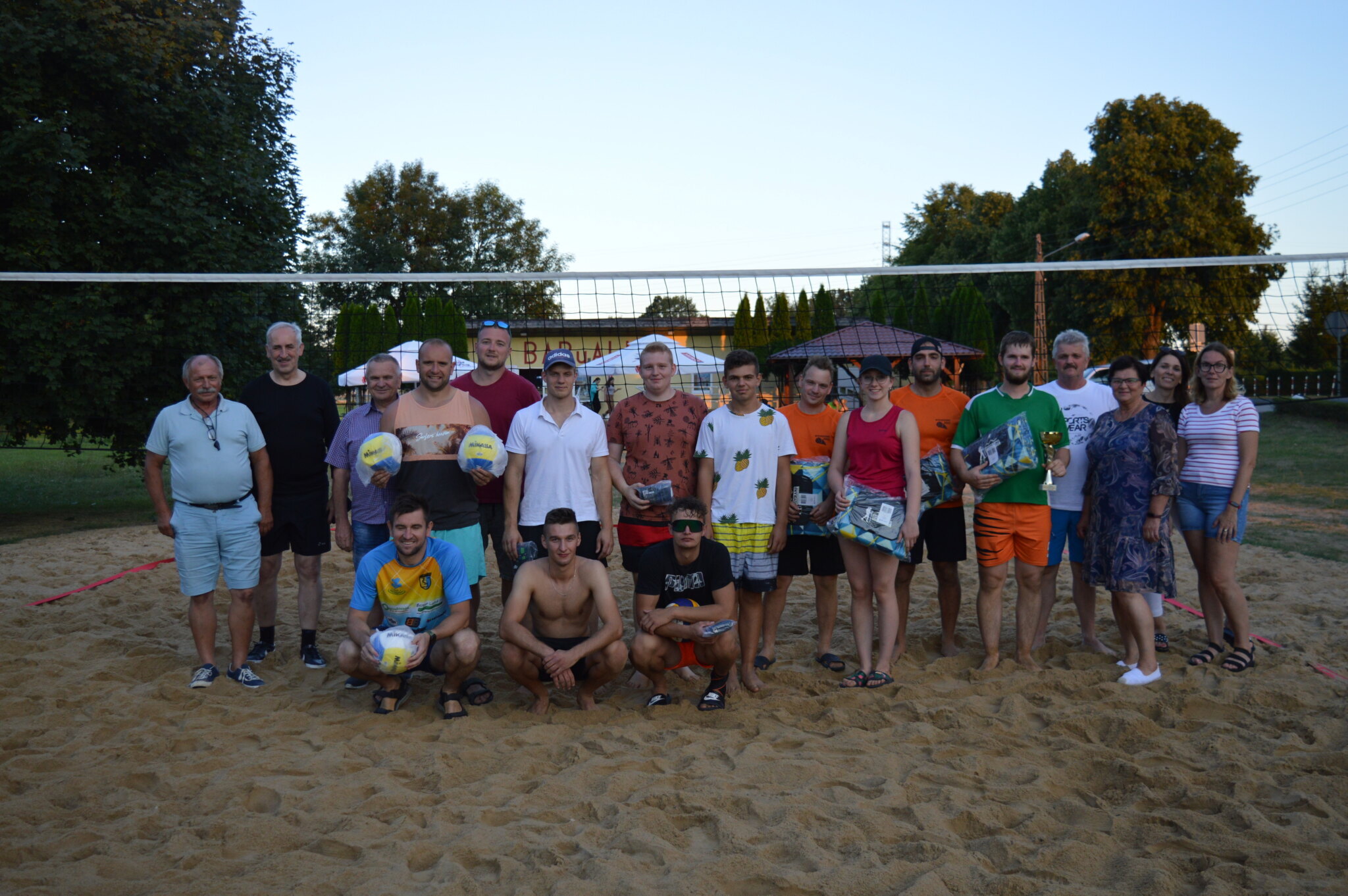 Zdjęcie grupowe uczestników Turnieju z organizatorami oraz Wójtem Gminy. Zdjęcie wykonano na polu do siatkówki plażowej. Zawodnicy trzymają w rękach nagrody oraz puchar.&nbsp;