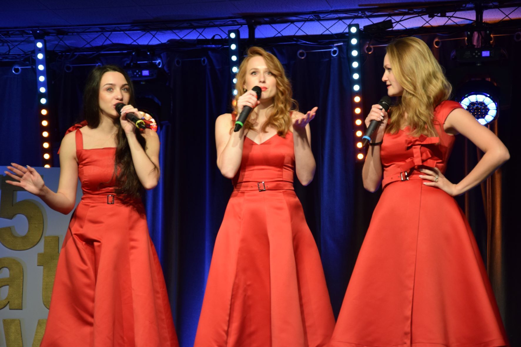 Ujęcie podczas występu. 
Na scenie trzy kobiety z mikrofonem w czerwonych sukienkach&nbsp;