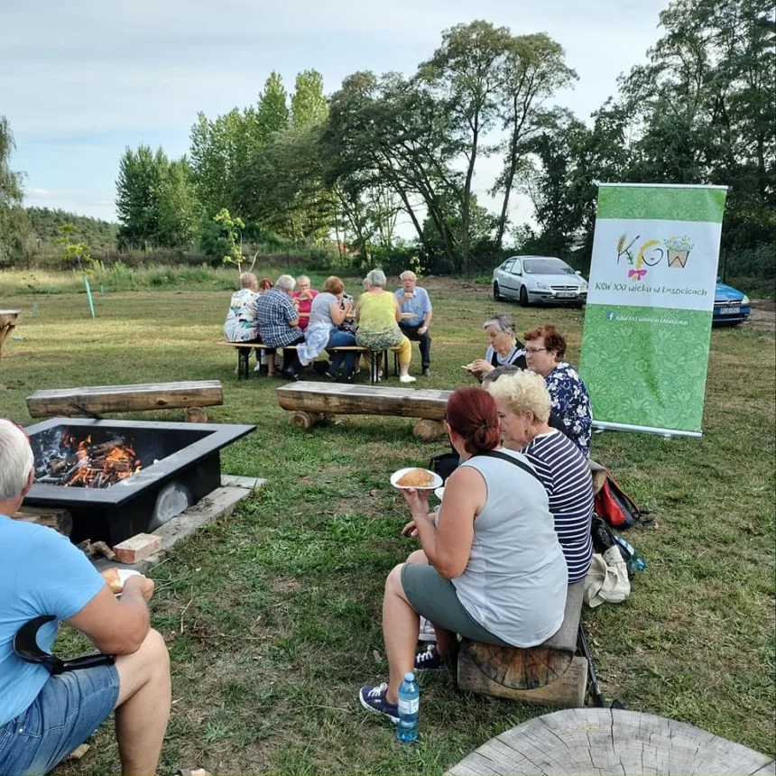 Ujęcie podczas grillowania. Seniorzy siedzą na ławkach i jedzą upieczone kiełbaski