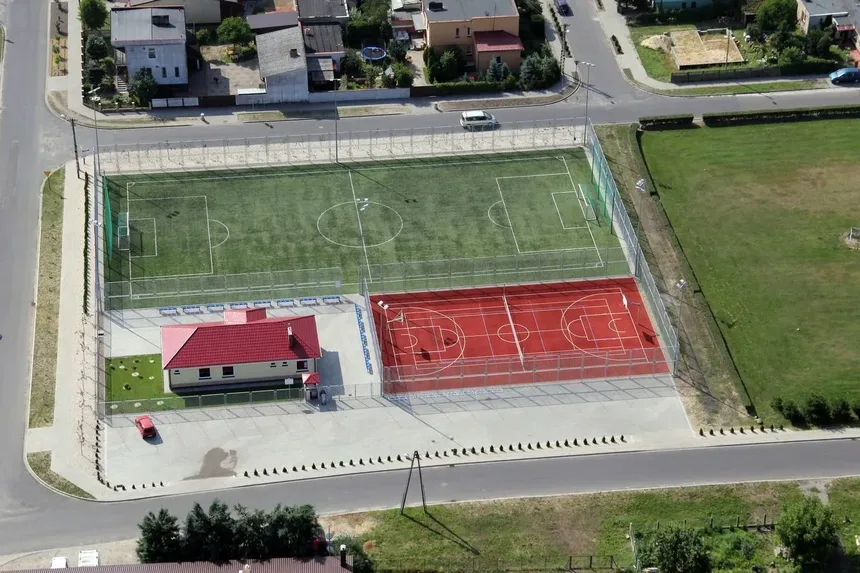 Zdjęcie z lotu ptaka orlika w Święciechowie. Na zdjęciu widać boisko do siatkówki i piłki nożnej oraz pomieszczenie gospodarcze. W oddali widać budynki jednorodzinne