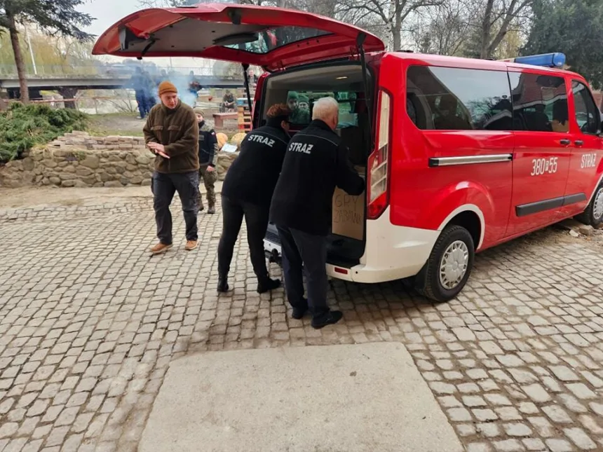 Osoby w mundurach strażackich zajmujące się ładunkiem w czerwonym pojeździe strażackim na placu wyłożonym kostką brukową, w tle widoczna jest zieleń i dym.