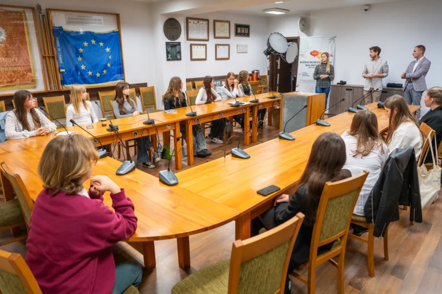 Spotkanie grupy młodych ludzi w sali konferencyjnej z drewnianym stołem, na którym znajdują się mikrofony. Na ścianie wisi flaga Unii Europejskiej. Uczestnicy skupieni na mówiącej osobie, która prezentuje informacje. W tle widoczne są dokumenty oraz sprzęt multimedialny.