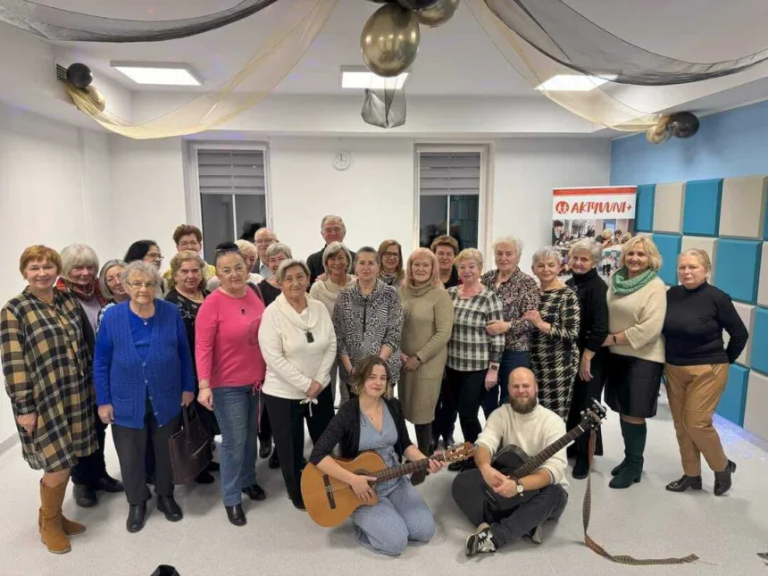 Grupa kobiet w różnym wieku stoi w pomieszczeniu, niektóre z nich trzymają instrumenty muzyczne. W tle dekoracje w postaci balonów i zasłon. Na ścianie widoczny banner z napisem "AKTYWNI+".