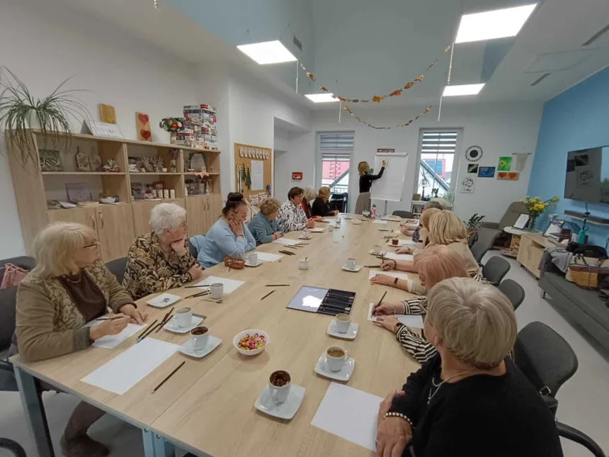 Grupa kobiet siedzących przy długim stole podczas warsztatów, z notatkami i filiżankami kawy, a jedna osoba prowadzi prezentację przy tablicy flipchart. W tle widoczne dekoracje na ścianach.