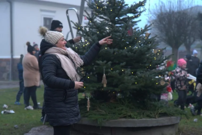 Kobieta w czarnej kurtce i beżowej czapce z pomponem wskazuje na dekoracje na choince, podczas gdy w tle widać innych ludzi bawiących się w parku.