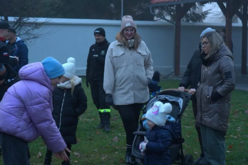 Grupa osób na zewnątrz, w tym dzieci bawiące się i dorosłych rozmawiających, w otoczeniu zielonej trawy i szarego nieba.