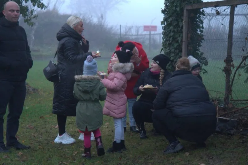 Grupa ludzi stoi i siedzi na trawie w mglistym otoczeniu, rozmawiając i jedząc ciasto. W tle widoczne jest ogrodzenie porośnięte bluszczem.