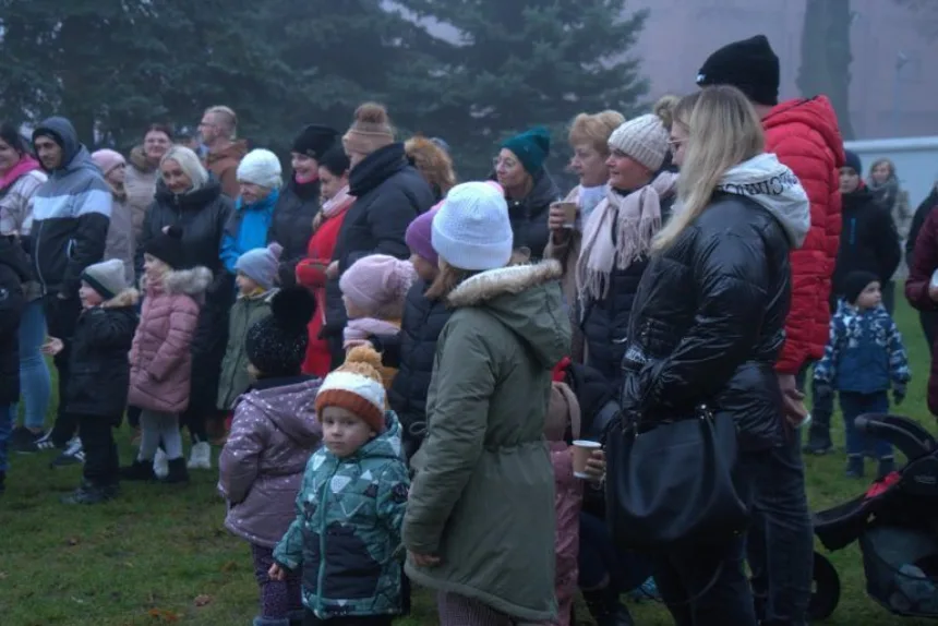 Grupa ludzi, w tym dzieci, stoi na zewnątrz w mglistej atmosferze, ubrani w ciepłe kurtki i czapki. Niektórzy trzymają kubki w rękach, a w tle widać drzewa.