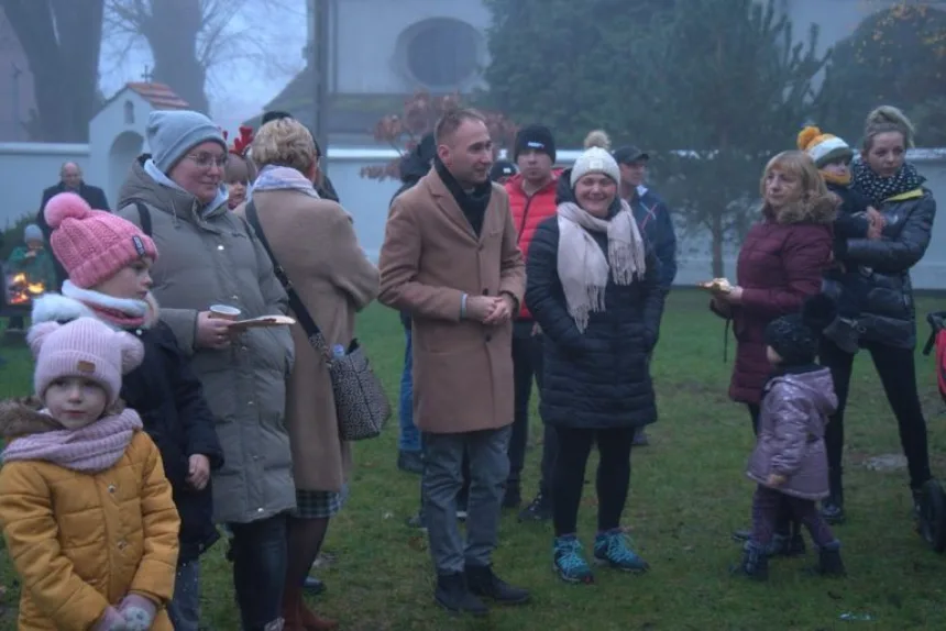 Grupa ludzi stoi na zewnątrz w parku, w otoczeniu mgły. Niektórzy trzymają kubki, a na tle widoczne są ognisko oraz budynki. Osoby są ubrane w ciepłe ubrania, a wśród nich są dzieci.