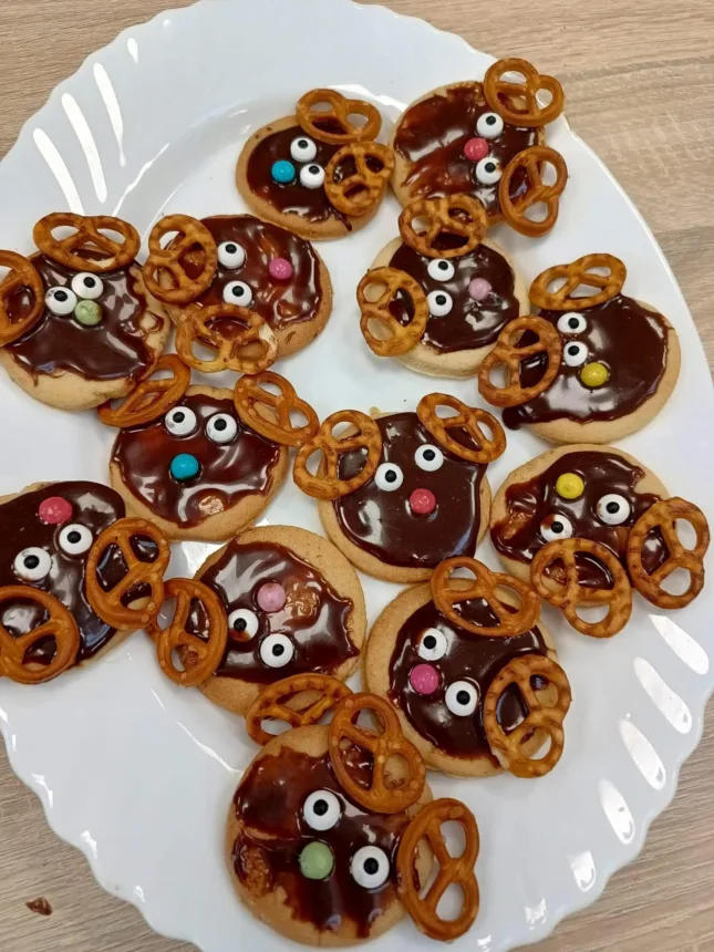 Na białym talerzu znajduje się kilka ciastek w kształcie głów zwierząt, pokrytych ciemną czekoladą. Ciasteczka mają na sobie pretzle jako uszy oraz kolorowe cukierki jako oczy i nosy.