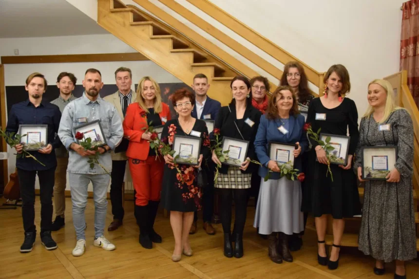 Grupa osób stoi na schodach, każda trzyma dyplom i różę. W tle widoczna jasna ściana i drewniane schody. Osoby są ubrane w różnorodne stylizacje, tworząc przyjazną atmosferę.