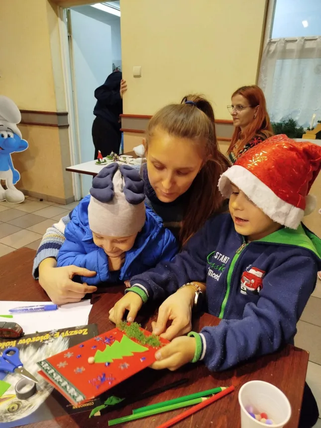 Dzieci bawią się razem przy stole, tworząc kartkę świąteczną. Jedno z dzieci trzyma w rękach czerwoną kartkę z ozdobami w kształcie choinki, a drugie śmieje się, patrząc na nią. W tle widać kobietę z rudymi włosami, a obok niej stoi postać w niebieskim kostiumie. Na stole znajdują się różne materiały do rękodzieła, takie jak nożyczki, klej i kolorowe pisaki.