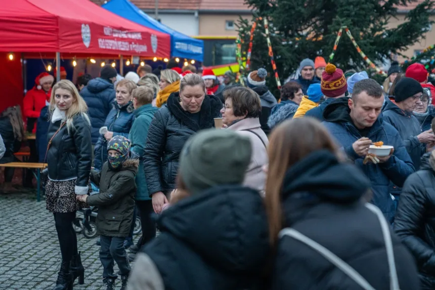 Na zdjęciu widać tłum ludzi zbierających się wokół kolorowych straganów na jarmarku, gdzie niektórzy trzymają jedzenie i napoje. Wśród zgromadzonych osób widać kobietę w skórzanej kurtce oraz dziecko w zimowej odzieży. W tle znajdują się budki z czerwonymi i niebieskimi daszkami oraz dekoracje bożonarodzeniowe. Atmosfera jest radosna i tłumna.