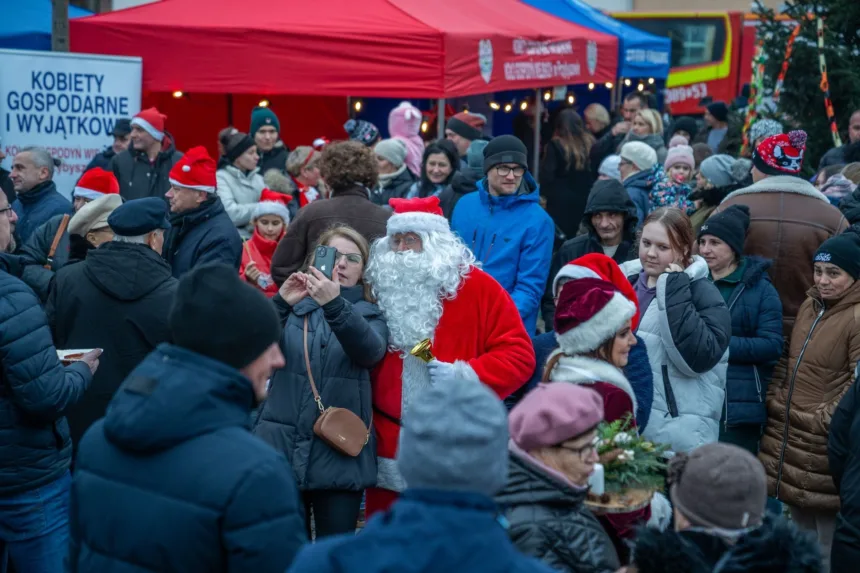 Tłum ludzi na jarmarku bożonarodzeniowym, wśród których wyróżnia się Święty Mikołaj pozujący do selfie z kobietą, w tle stoiska z dekoracjami i świątecznymi czapeczkami.