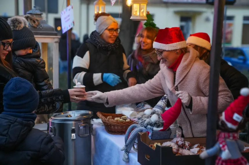 Grupa osób uczestniczących w kiermaszu, gdzie jedna z kobiet w czerwonej czapce podaje porcję jedzenia, a inne osoby w różnorodnych ubraniach rozmawiają i obserwują sytuację. W tle widać lampiony i stół z jedzeniem.