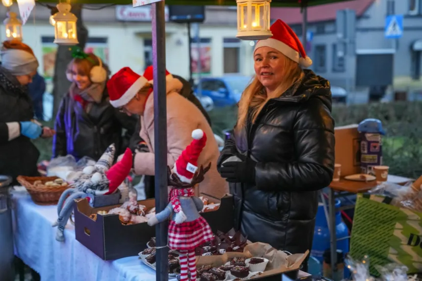 Kobieta w czarnej kurtce, z długimi blond włosami, stoi przy straganie na jarmarku świątecznym, w czerwonej czapce Mikołaja, podczas gdy inne osoby w zimowych ubraniach sprzedają wypieki i ozdoby. W tle widać lampiony i kolorowe reklamy.