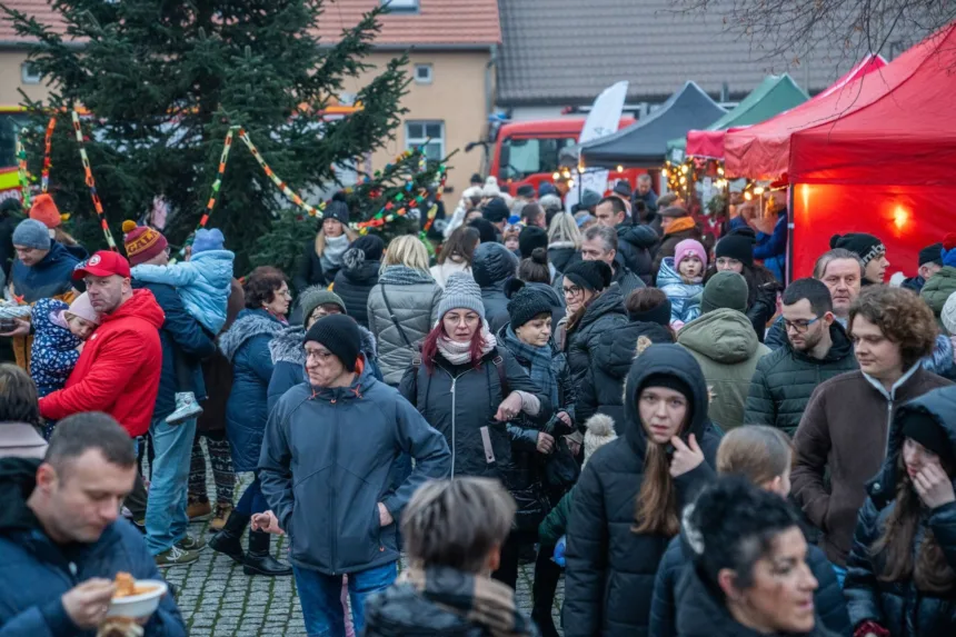 Tłum ludzi na festynie lub jarmarku, wśród którego widać stoisko z czerwonym dachem oraz choinkę ozdobioną światełkami; niektórzy trzymają jedzenie, a wielu z uczestników ma na sobie ciepłe kurtki i czapki.