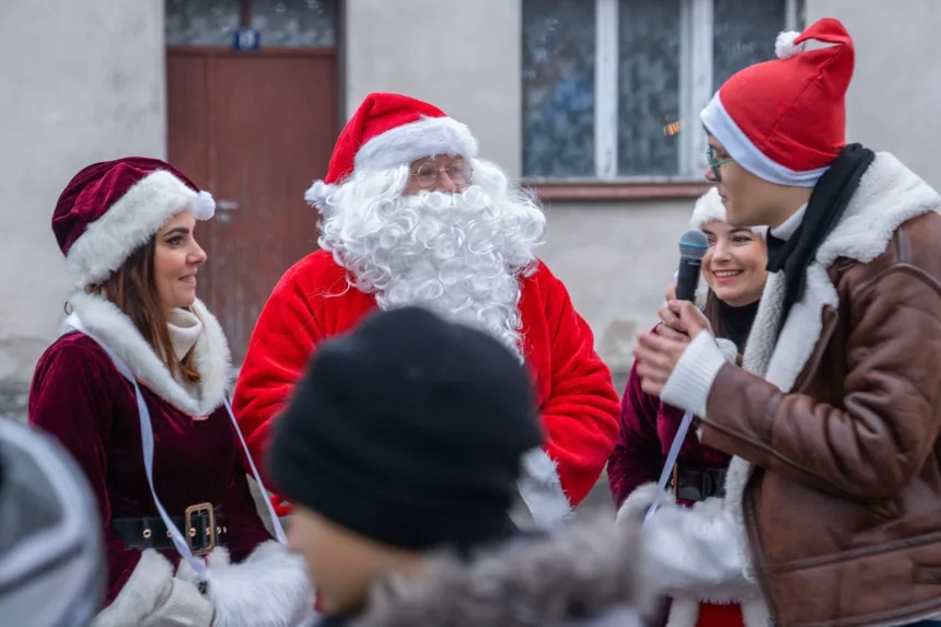 Na zdjęciu widać Mikołaja z długą, białą brodą w czerwonym stroju, który rozmawia z dwiema kobietami ubranymi w czerwone kostiumy świąteczne z białymi akcentami oraz mężczyzną trzymającym mikrofon. W tle znajduje się budynek z zamkniętymi oknami.