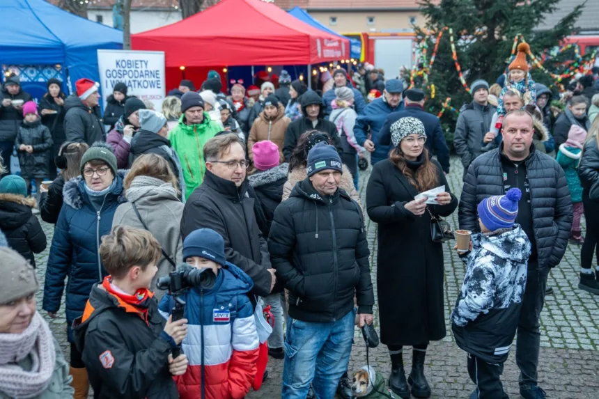 Ludzie zgromadzeni na jarmarku, w tle kolorowe namioty, dekoracje świąteczne, dzieci bawiące się z rodzicami, piesek na smyczy.